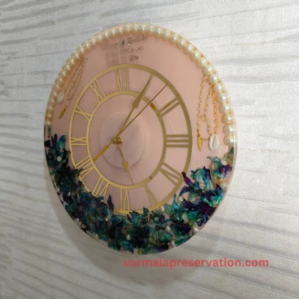 Capture timeless love with Pink & Blue Theme Resin Varmala Preserved Wall Clock featuring Roman Numerals, Couple Name, Hashtag, and Wedding Date. Preserve your cherished varmala in epoxy resin, adorned with Half Pearls & Half Floral Decor (12 Inch) for everlasting memories.